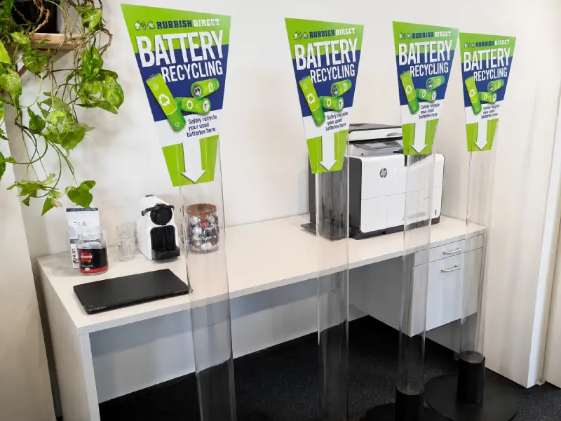 Office Corner with Four Clear Acrylic Barriers and Green aposbattery Recyclingapos Banners over a Desk with a Printer Plant and Laptop