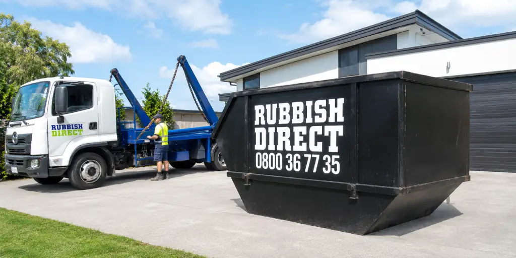 Rubbish Direct Truck with a Blue Crane Trailer and a Worker in a Safety Vest Beside a Large Black Dumpster in a Driveway Outside a Modern House