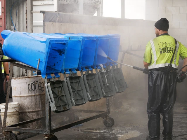 Worker in a High visibility Vest Rinses Blue Wheelie Bins on a Rack in an Industrial Wash Area with a Hose