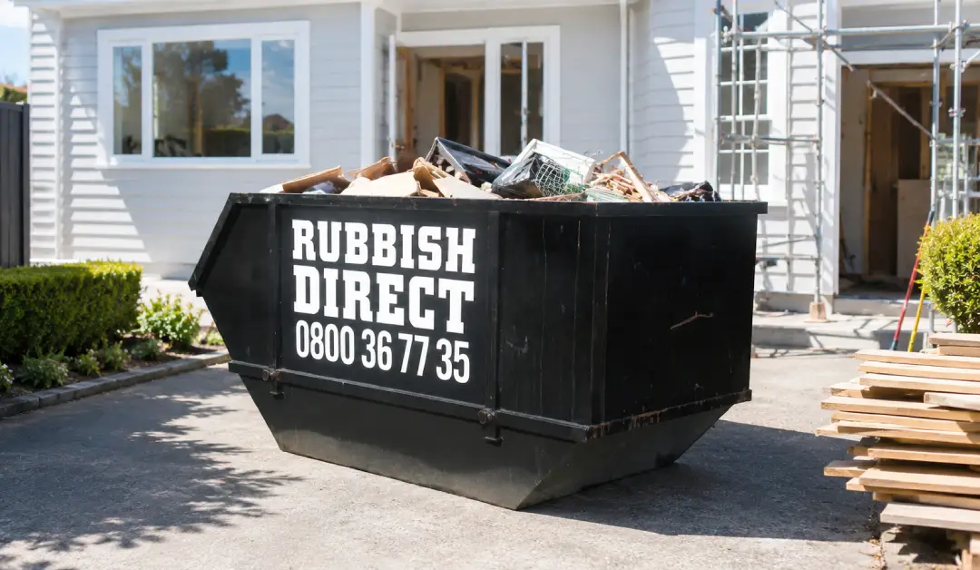 Black Skip Filled with Debris Sits on a Driveway in Front of a Partially Built House with Scaffolding and New Timber Nearby and the Label Rubbish Direct Visible on the Side