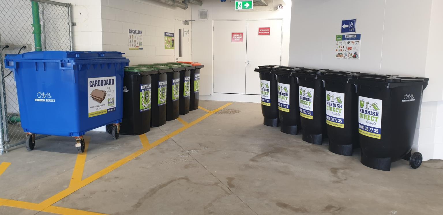 Blue Recycling Bin and a Row of Black Waste Bins Along a Hallway with Yellow Floor Lines and an Exit Sign Above the Doors