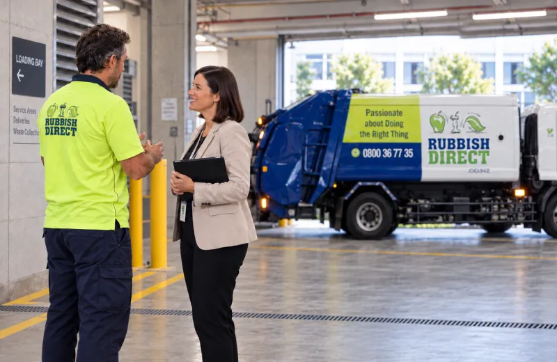 Worker in a Neon Uniform Talks with a Woman Holding a Clipboard Inside a Waste Depot a Rubbish Direct Truck in the Background