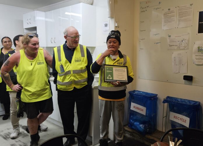 Celebrating incredible years with resa penelijpg | Rubbish Direct Group of People in High visibility Vests Indoors a Woman Holds a Framed Certificate While Another Speaks to the Group in a Kitchen Area with Recycling Bins Nearby