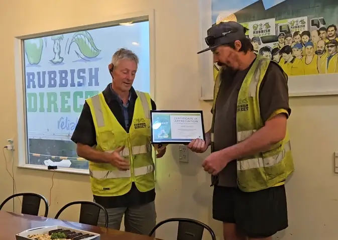 Celebrating our workshop hero mark dunkleywebp | Rubbish Direct Two Men in Yellow Safety Vests Exchange a Certificate of Appreciation Inside a Meeting Room with rubbish Direct Sign Visible Outside the Window